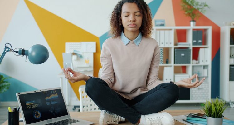 Young African American girl is sitting in lotus pose on office table enjoying relaxation and meditation. Job, stress management and break at work concept.